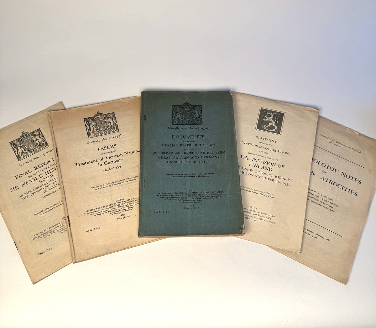 Five vintage printed pamphlets or booklets arranged in a fan shape on a white surface. The center booklet has a muted blue-green cover with black text and crest, while the surrounding four booklets have aged tan paper covers with black printed text and decorative emblems.