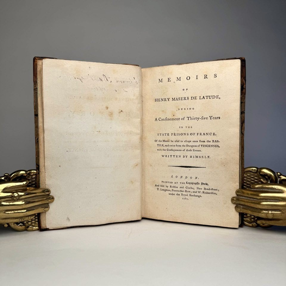 An open antique book titled 'Memoirs' by Henry Meusnier de Latude, showing aged, yellowed pages with printed text, displayed on a white surface with decorative gold-toned bookends on both sides against a gray background.